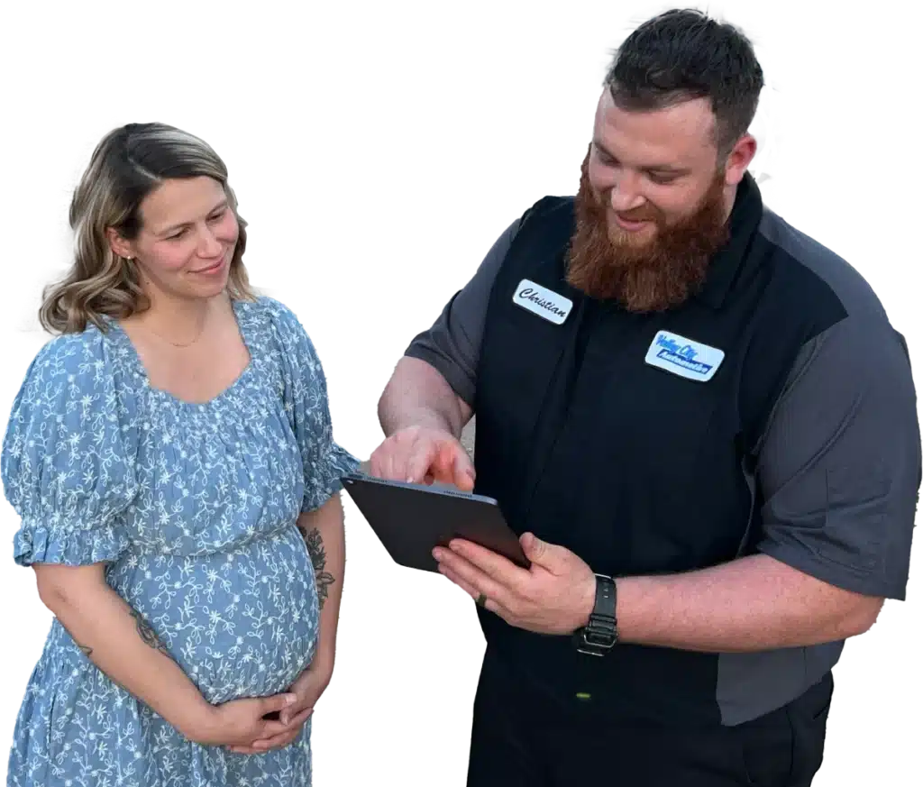 A man wearing a dark uniform and name badge from Valley City Automotive shows a tablet to a smiling woman in a blue floral dress. The woman holds her hands together in front of her. Both appear engaged and happy, discussing auto repair options.