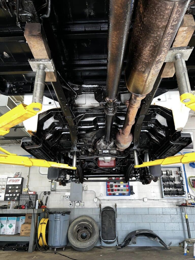 View of a car's undercarriage from below while it is lifted on a hydraulic car lift in an auto repair shop. The lift's yellow arms are visible, alongside aftermarket wheels and various auto accessories scattered around the shop floor amidst tools, tires, and equipment.