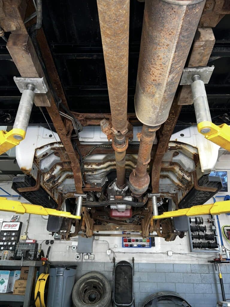 View of the underside of a vehicle raised on a hydraulic lift in an automotive repair shop. Visible components include rusty exhaust pipes, drive shaft, and suspension system. Various tools, equipment, tires, and parts are scattered around the brightly lit shop, including aftermarket wheels and lift kits.